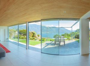 Distorted interior view so show the view outside too. Plain light-toned ceramic floor tiles and side walls. Panoramic view of Lake Maggiore and mountains beyond. Acoustically grooved wooden ceiling in this holiday home. House plans.
